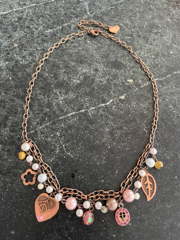 A handmade copper chain necklace with pink and white beads, a copper leaf charm, a pink heart charm, and a clover charm, displayed on a dark textured slate surface.