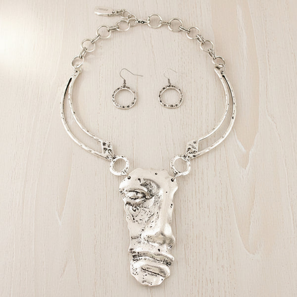 A silver-toned jewelry set featuring a textured abstract pendant necklace and matching circular drop earrings, displayed on a neutral gray background.