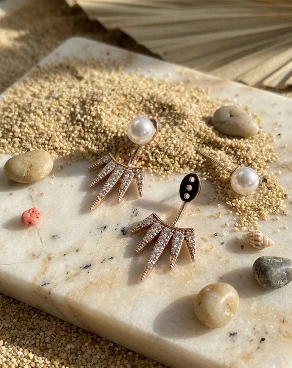 Pair of earrings with pearl-like stones on a textured surface with stones and sand.