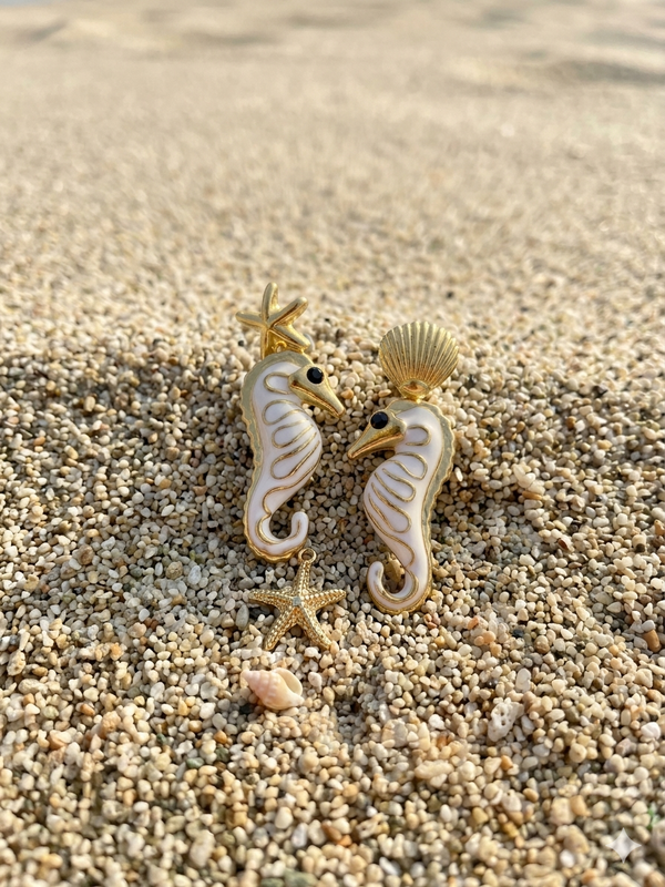 Pair of seahorse-shaped earrings on sand with starfish and shells.