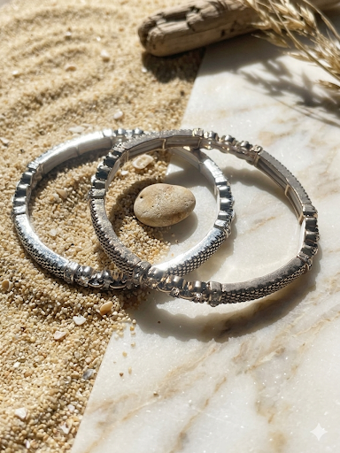 Two silver bangles on a stone surface with sand and driftwood in the background