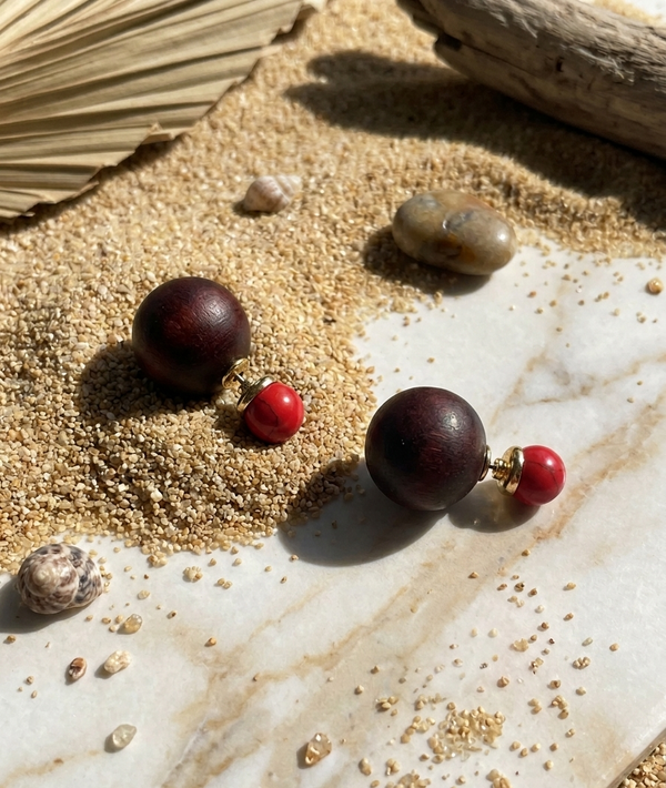 Pair of earrings with wooden and red beads on a textured surface with sand and driftwood.