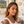 Load image into Gallery viewer, Woman with wavy hair and earrings standing outdoors near a stone wall.

