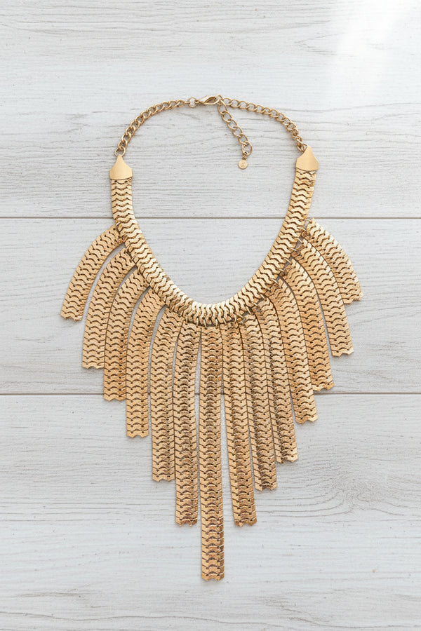 A gold-toned statement necklace with a textured, scale-like fringe design, displayed on a dark slate surface.