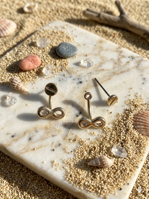 Gold infinity-shaped earrings on a marble surface with seashells and sand.