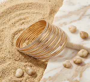 Stack of gold bangles with diamond accents on a sandy surface with stones.