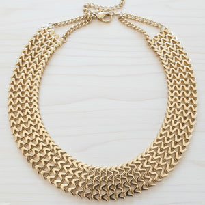 A wide, multi-strand gold chain necklace with a geometric, textured chevron pattern, displayed on a dark brown wooden surface.