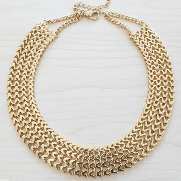A wide, multi-strand gold chain necklace with a geometric, textured chevron pattern, displayed on a dark brown wooden surface.