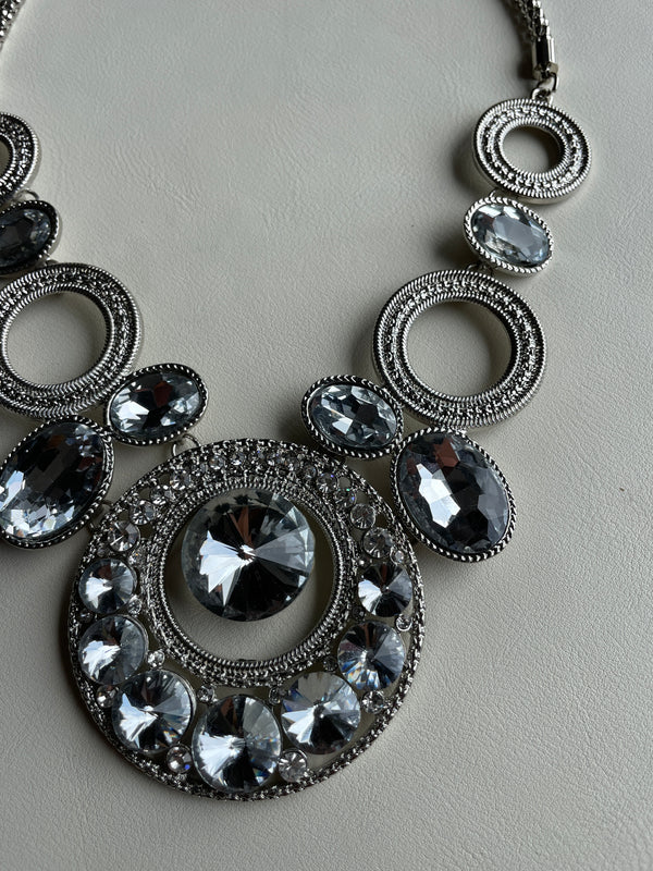 A close-up of a statement necklace featuring silver-toned filigree rings and oval links set with clear faceted crystals on a light gray textured background.