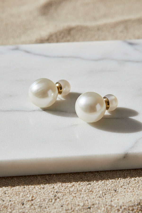 A pair of drop earrings with large, milky white marbled beads connected by small gold spacers to smaller, pale pinkish-white beads, displayed on a neutral light gray background.