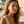 Charger l'image dans la galerie, Woman with earrings standing in front of a scenic background with buildings and palm trees.

