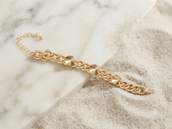 A gold chain bracelet with textured, faceted disc links, styled diagonally across a split background of white marble and light beige sand.