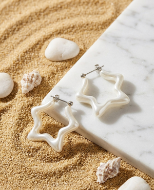 Star-shaped earrings on a marble surface with sand and shells around