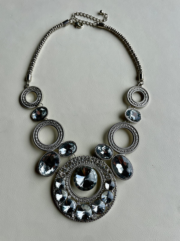 A silver-toned statement necklace featuring multiple circular and oval links, some adorned with clear faceted rhinestones, laid flat on a light gray background.