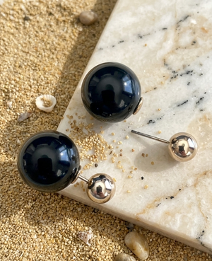 Black spherical earrings with gold posts on a marble surface with sand and small stones.