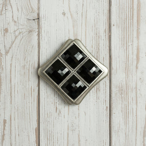 A vintage-style diamond-shaped brooch featuring four inset black faceted gemstones divided by a silver cross, set within a textured silver border on a white background.