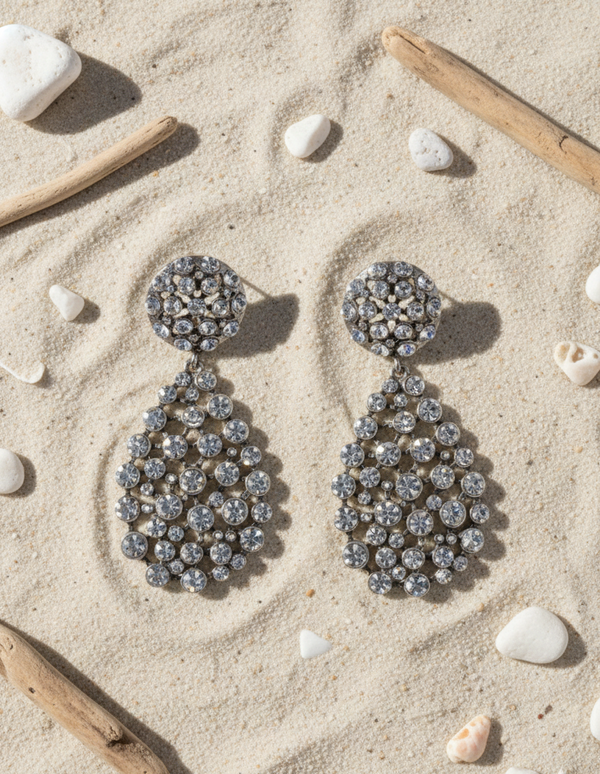 Pair of sparkling earrings on a sandy surface with driftwood and shells.