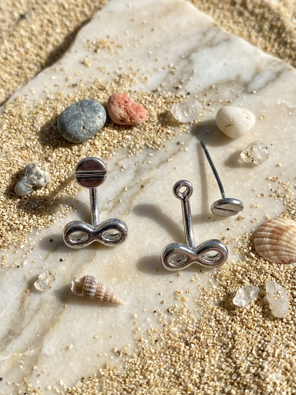 Silver infinity symbol earrings on a stone surface with seashells and sand.
