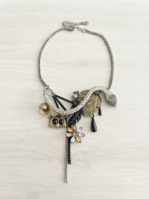 A silver-toned statement necklace featuring a snake, leaf, owl, dragonfly, and teardrop charms, displayed on a light wood background.