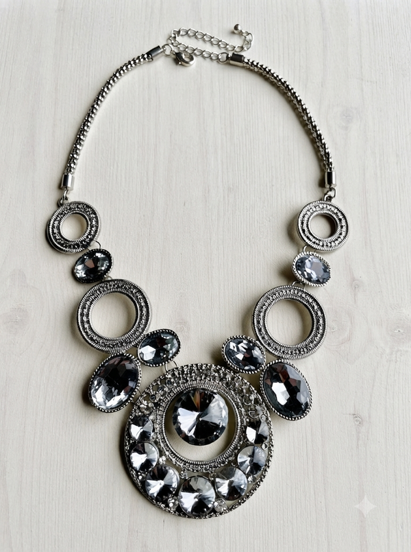 A statement bib necklace featuring a silver-tone chain and multiple circular and oval links adorned with faceted clear and dark gray rhinestones, displayed on a light wood background.