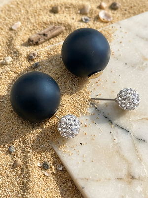 Black spherical earrings with silver studs on a sandy surface