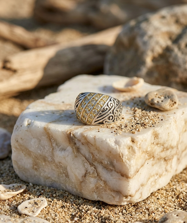 Gold ring with intricate design on a stone surface with sand and shells around