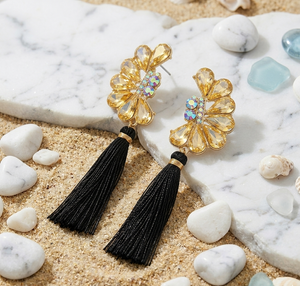 Gold floral earrings with black tassels on a marble surface with stones and shells.