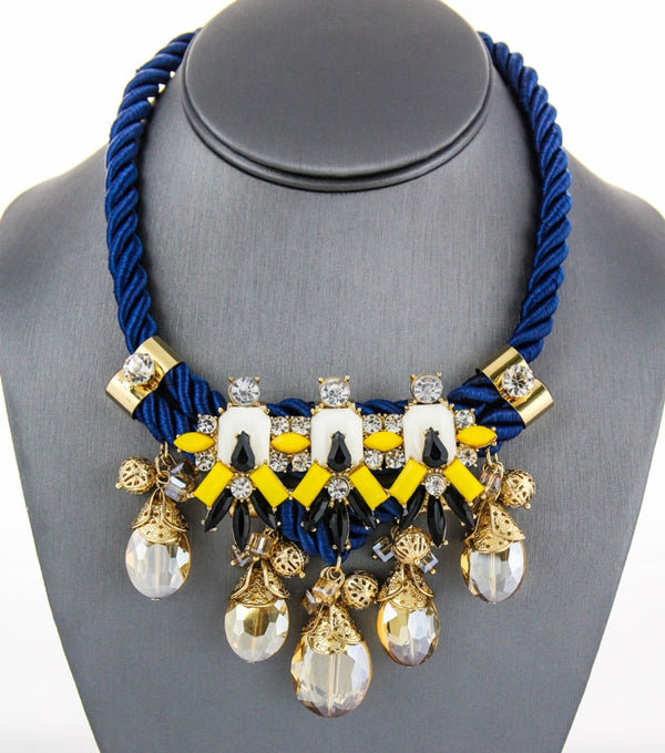 A statement bib necklace displayed on a gray mannequin bust. The necklace features a thick royal blue braided cord base with gold-tone metal accents, white and yellow geometric beads, and dangling clear faceted crystal drops.
