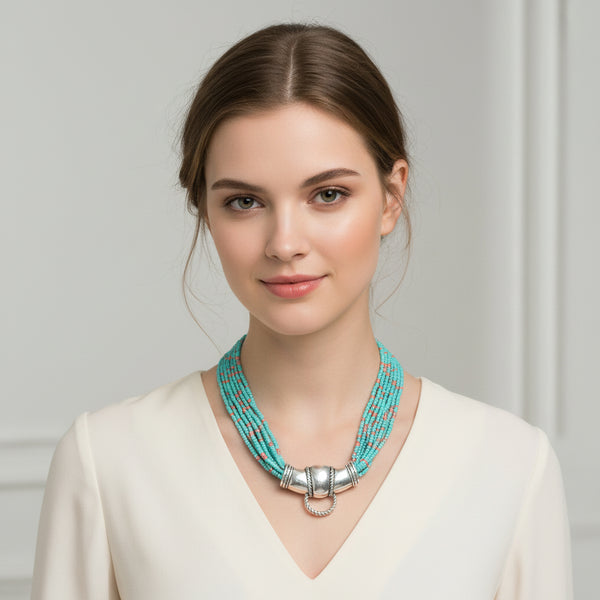 A multi-strand turquoise beaded necklace with small coral accents and a polished silver centerpiece, displayed on a textured gray jewelry bust.