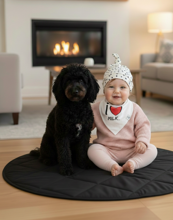 Child in a pink outfit with a heart-themed bib sitting next to a black dog on a black mat in a living room.