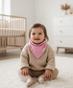 Child wearing a beige sweater and pink bib sitting on a white carpet in a nursery.