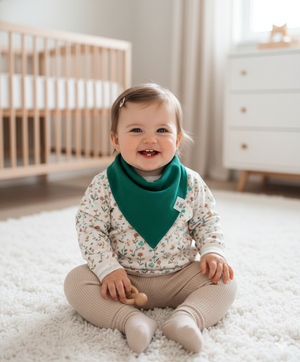 Baby sitting on a white rug in a nursery with a crib and dresser in the background.
