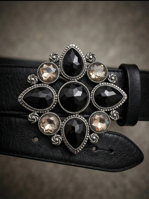 A close-up of a black leather belt featuring an ornate silver-tone buckle adorned with faceted black and smoky-tinted crystals on a textured gray background.