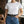 Charger l'image dans la galerie, A woman wearing a plain white short-sleeve crewneck t-shirt tucked into blue jeans with a black belt featuring a large decorative silver-toned buckle, standing in a loft-style room with brick walls and windows.
