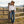 Load image into Gallery viewer, A woman stands outdoors in a rustic ranch setting, leaning against a wooden post. She is wearing a brown cowboy hat, a white long-sleeve button-up shirt, blue jeans, and brown boots, with a wooden fence and mountains in the background.
