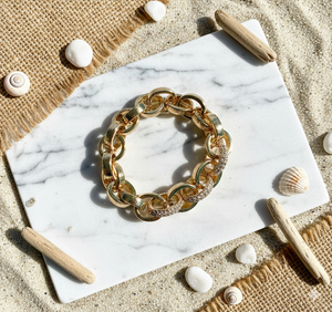 Gold chain bracelet on a marble surface with beach elements like shells and driftwood.