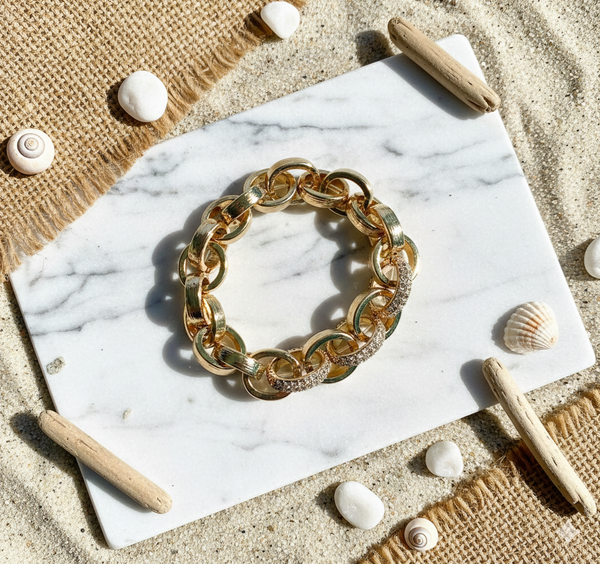 Gold chain bracelet on a marble surface with beach elements like shells and driftwood.