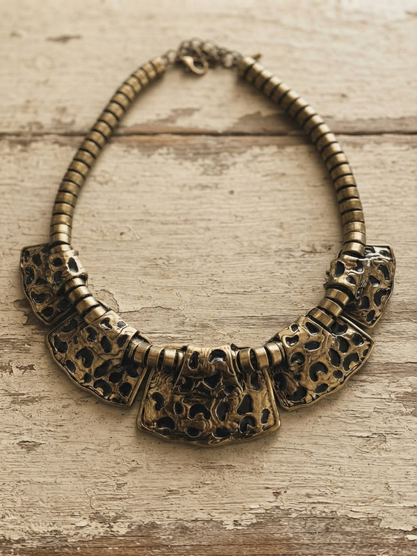 An antique bronze statement necklace featuring a beaded chain and three large, textured skull-shaped center plates, displayed on a rustic, weathered wood background.