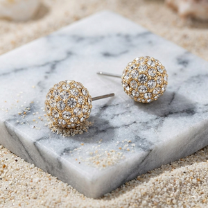 Gold and crystal stud earrings on a marble surface with sand around