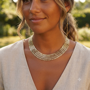 A close-up lifestyle shot of a woman wearing a chunky, woven gold chain necklace and matching drop earrings, set against a soft-focus outdoor background.