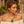 Load image into Gallery viewer, A woman wearing a chunky gold-toned statement necklace with a large reddish-brown stone pendant, matching drop earrings, and an off-the-shoulder cream top, photographed in a warm outdoor setting with blurred plants and stone architecture in the background.
