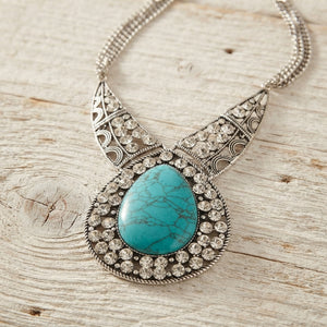 A silver filigree statement necklace featuring a large oval turquoise stone surrounded by clear rhinestones, displayed on a rustic light wood surface.
