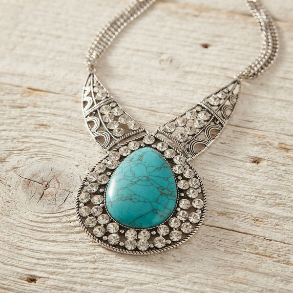 A silver filigree statement necklace featuring a large oval turquoise stone surrounded by clear rhinestones, displayed on a rustic light wood surface.