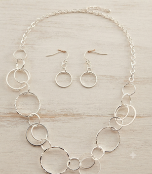 A silver jewelry set featuring a statement necklace and matching drop earrings, both adorned with textured and smooth interlocking circular rings, displayed on a light wood background.