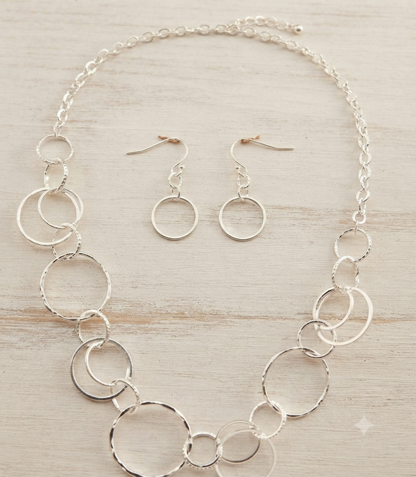 A silver jewelry set featuring a statement necklace and matching drop earrings, both adorned with textured and smooth interlocking circular rings, displayed on a light wood background.