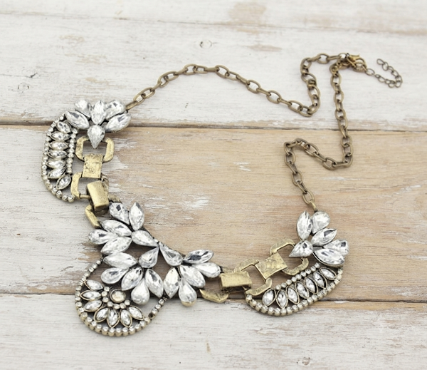 An ornate bib-style statement necklace with silver-toned teardrop rhinestones and gold-toned chain links, displayed on a light wood plank background.