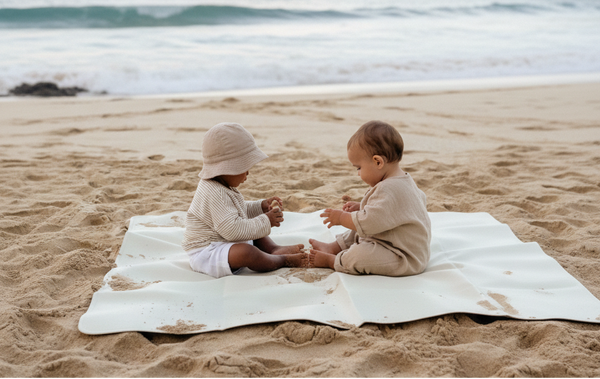Two children sitting on a blanket on a sandy beach with ocean and mountains in the background.