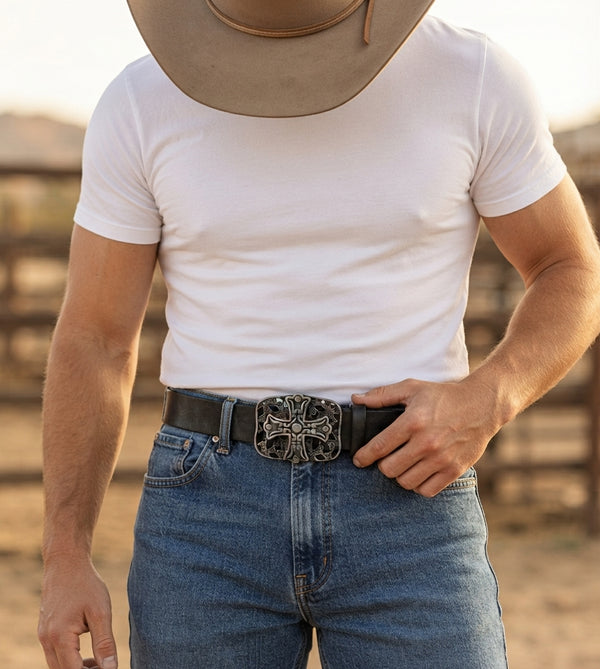 A man wearing a white crew-neck t-shirt, blue jeans, and a black leather belt with a large silver-toned decorative buckle, standing in a sunlit outdoor setting.