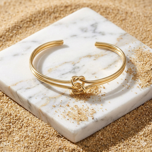 Gold bracelet with knot design on a marble surface with sand around