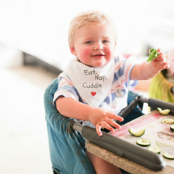 Child in a high chair with a bib reading 'Eat, Nap, Cuddle' holding a piece of broccoli.